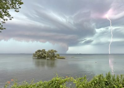 Looking out over Lake Ontario as a storm moves through, with a small island on the left, a large storm cloud above, and a lightning strike to the right, 2024 Photo Contest Best in Show winner, over 18, by Bridget Hughes