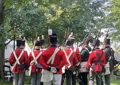 Portrait photo of the backs of reenactment soldiers during a briefing at the War of 1812 Grand Tactical event in July; 2024 Photo Contest winner in the Historic Sackets Harbor category, by Bob Smith (tied for first place)