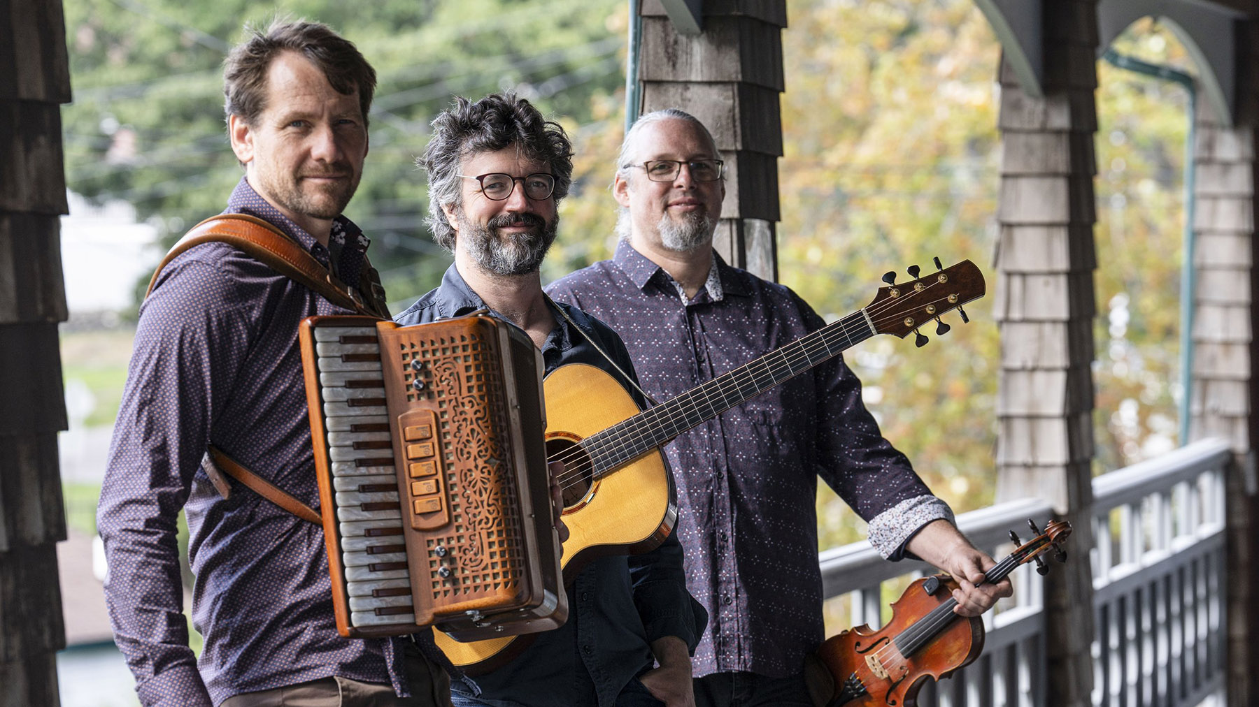 Publicity photo of the musical trio Genticorum, standing on an outdoor porch with their instruments