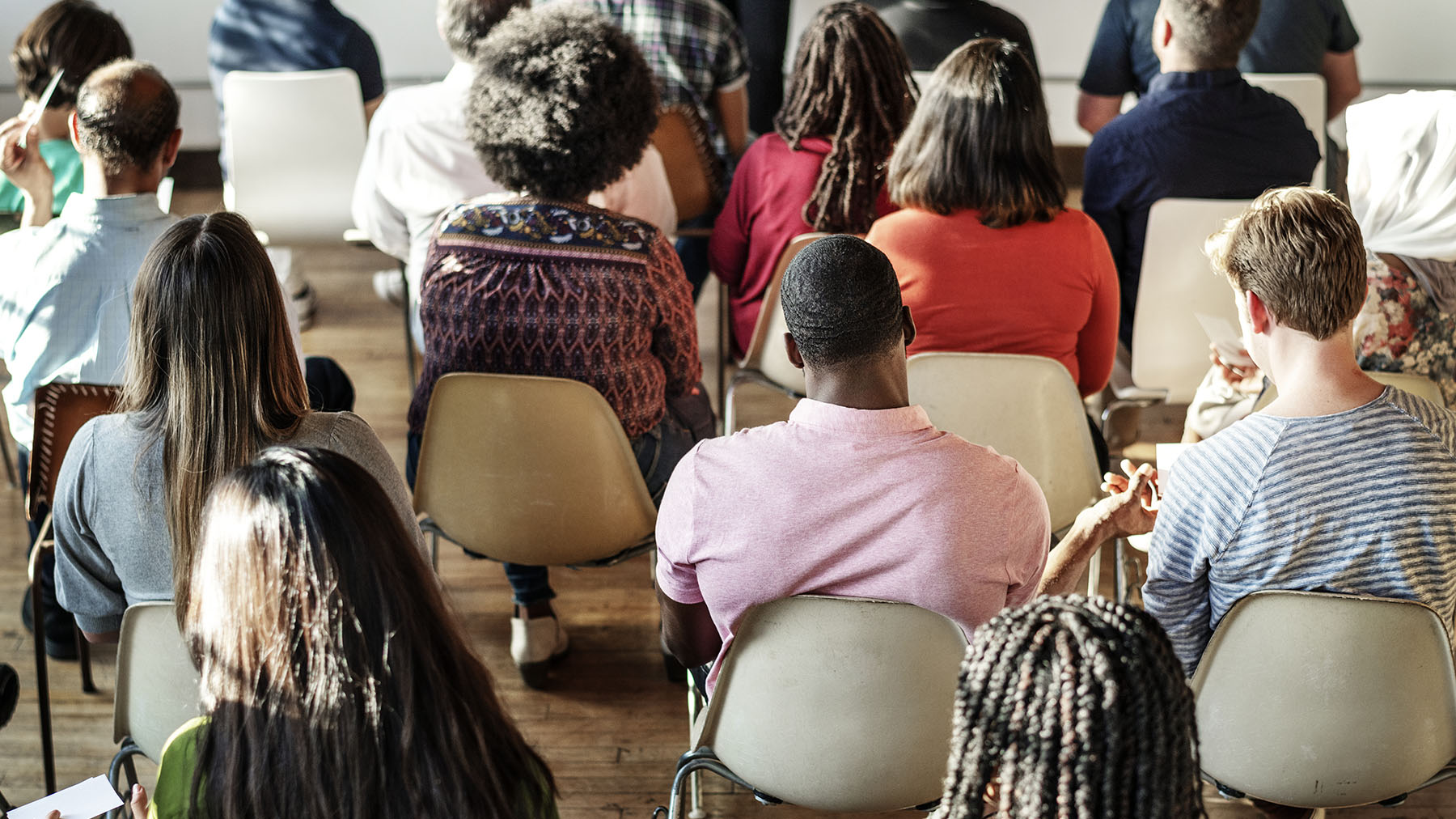 Color photo of the backs of a number of people sitting in chairs, attending a meeting (Image by RawPixel.com; copyright 2019)