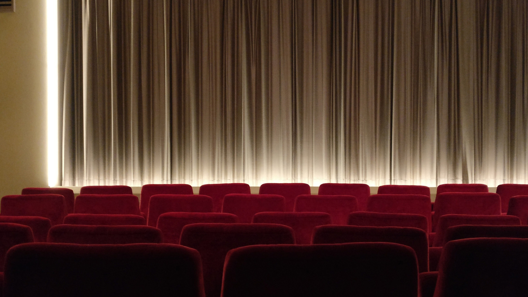 Stock image of a movie theater with lights on stage and empty red seats (Image by RawPixel.com)